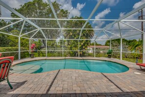 A covered swimming pool with a brick deck, surrounded by lush greenery and a metal frame structure.