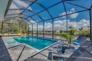 A covered outdoor swimming pool with lounge chairs, a laptop, and a bottle on a table. - Villa Southbreeze by Vacationhit (Cape Coral)