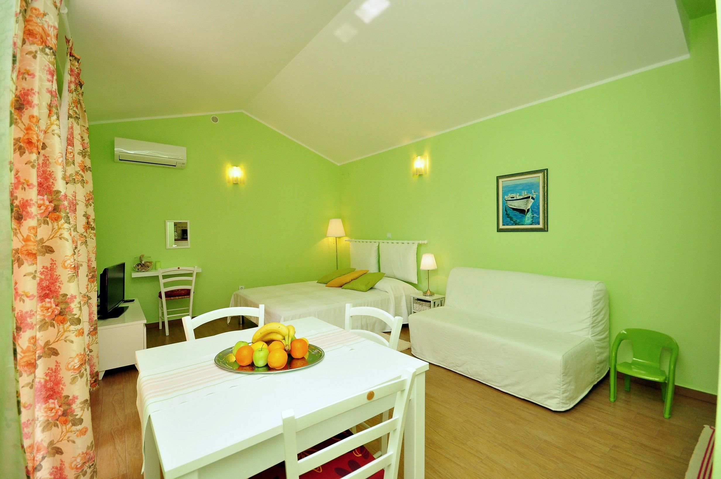 Una habitación con una cama, un escritorio, una silla, una mesa con fruta y otra silla verde.