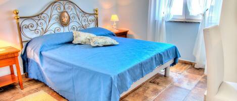 A neatly made bed with a blue bedspread and pillows, a wooden nightstand with a lamp, and a white chair.
