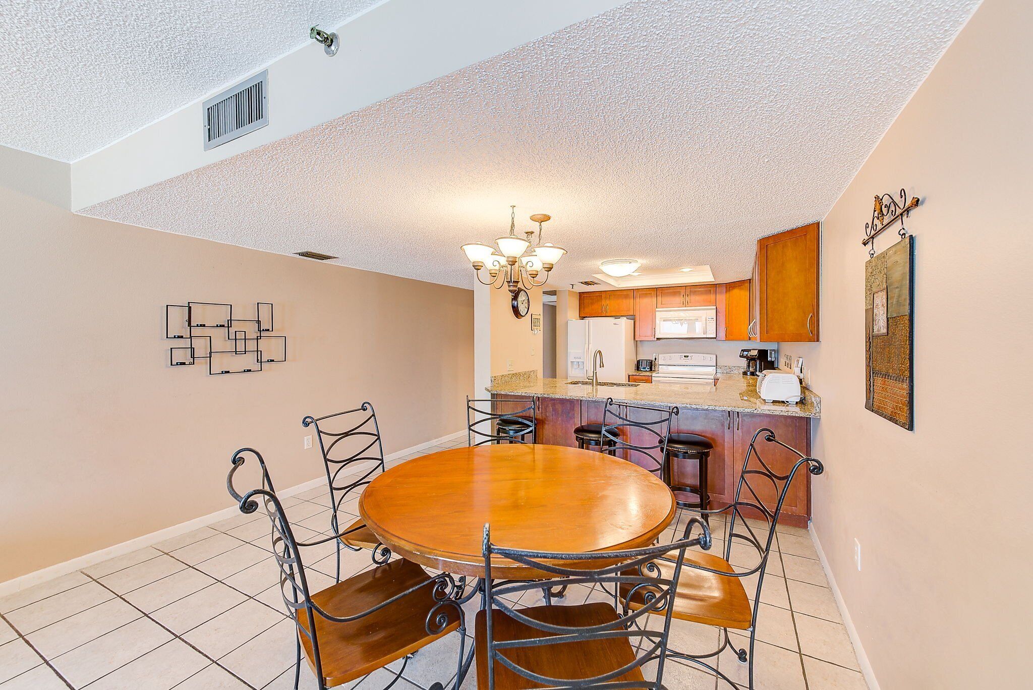 A dining area with a round wooden table and metal chairs, a chandelier, and a kitchen in the background.