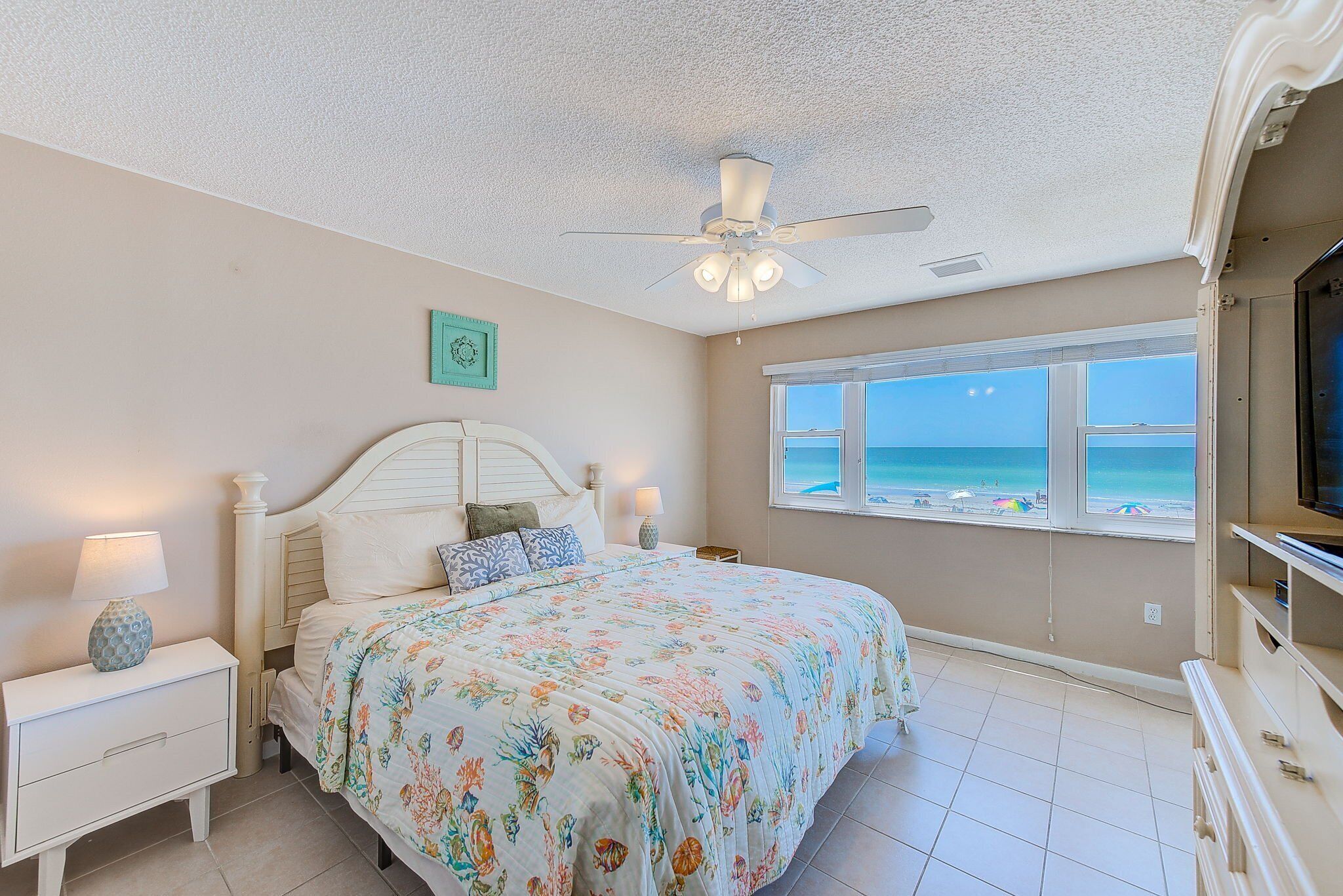 A bedroom with a bed, a ceiling fan, a nightstand with a lamp, a dresser, and a window with a view of the beach.