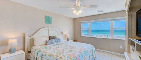 A bedroom with a bed, a ceiling fan, a nightstand with a lamp, a dresser, and a window with a view of the beach.