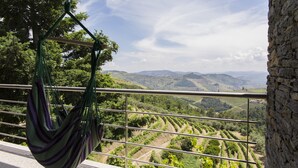 Un hamac est suspendu à une rambarde métallique, offrant une vue sur un vignoble avec des terrasses et des collines lointaines.