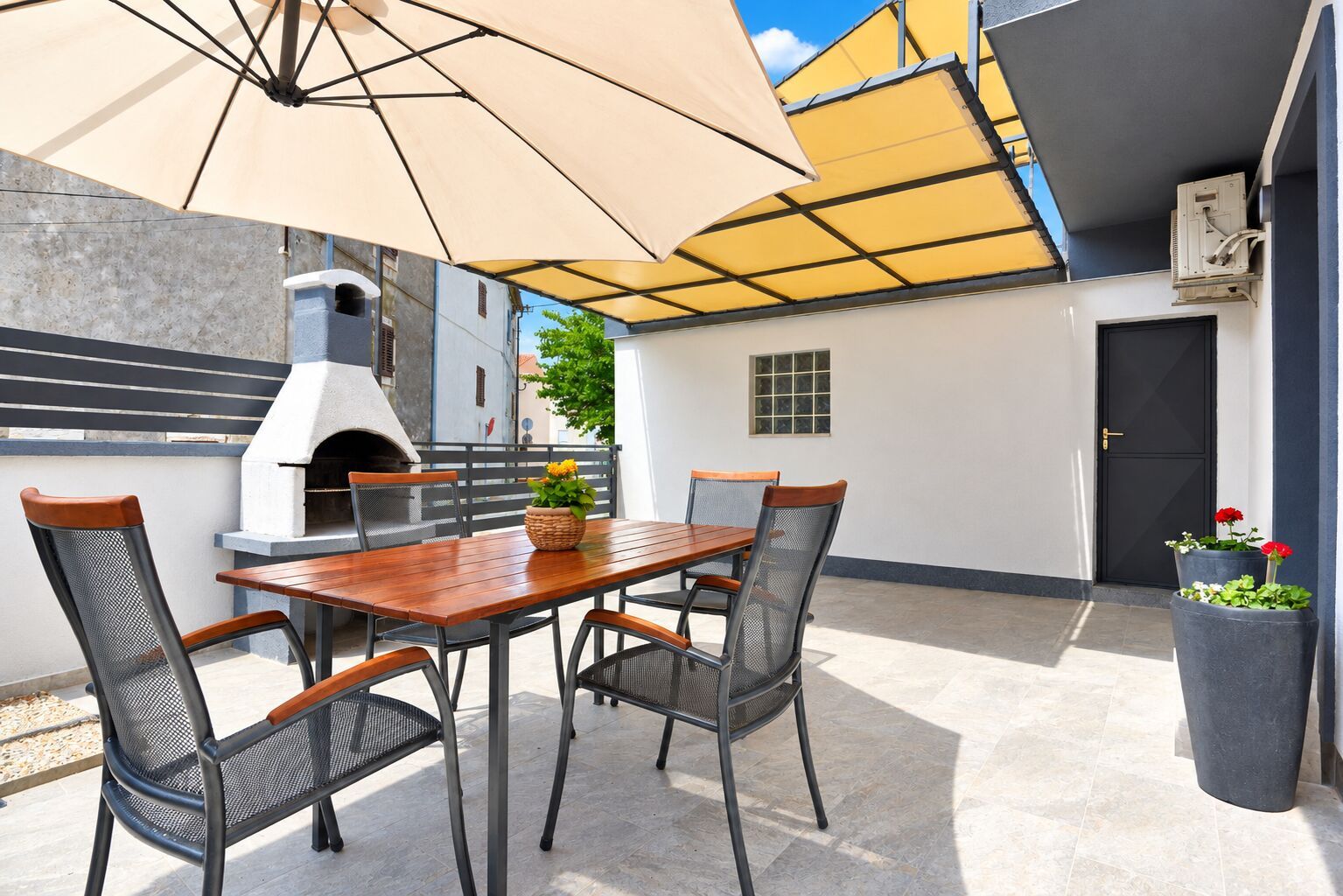 An outdoor patio with a wooden table, four chairs, a large umbrella, a pizza oven, and potted plants.