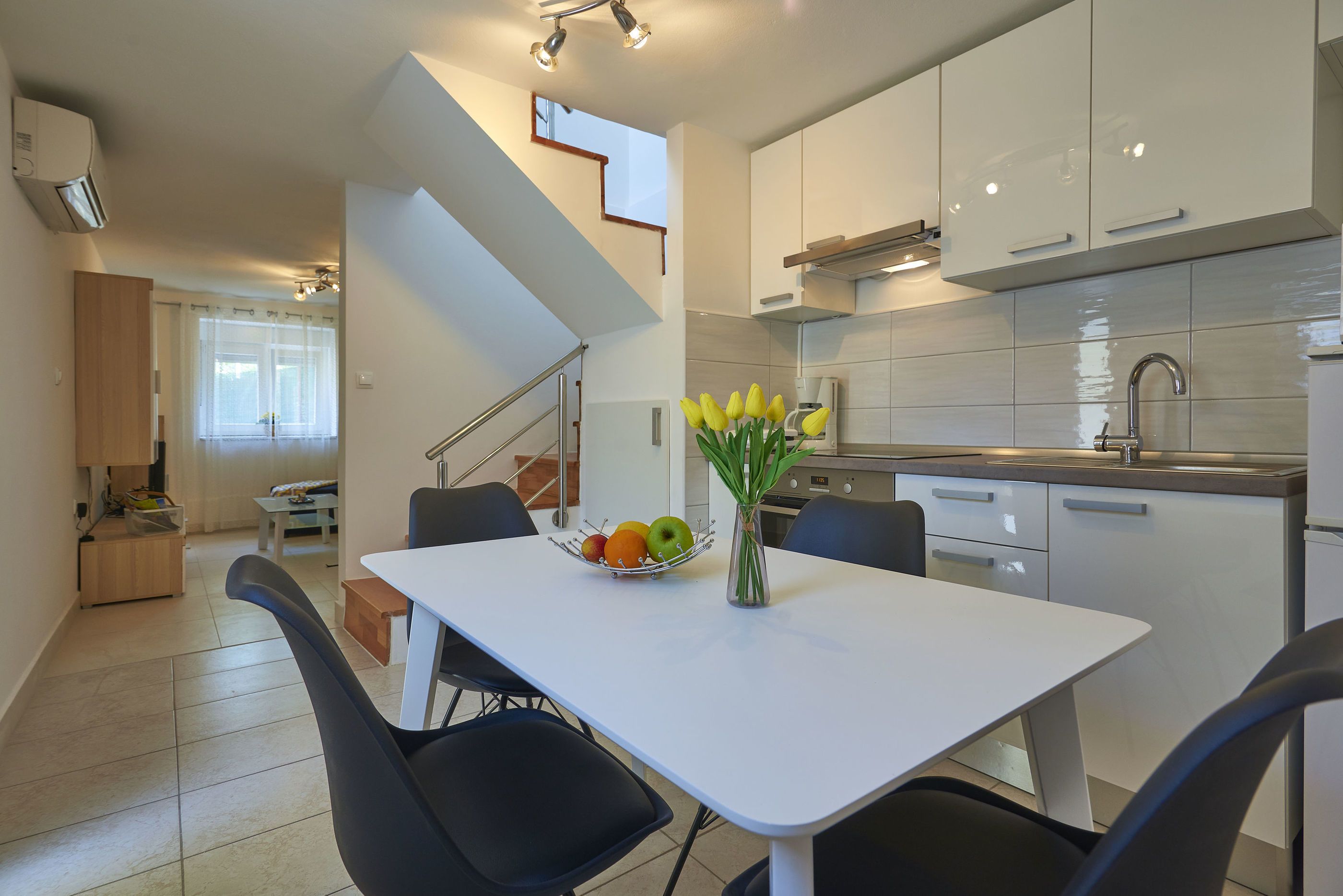 A modern kitchen with white cabinets, a dining table with black chairs, and a fruit bowl with apples and oranges.