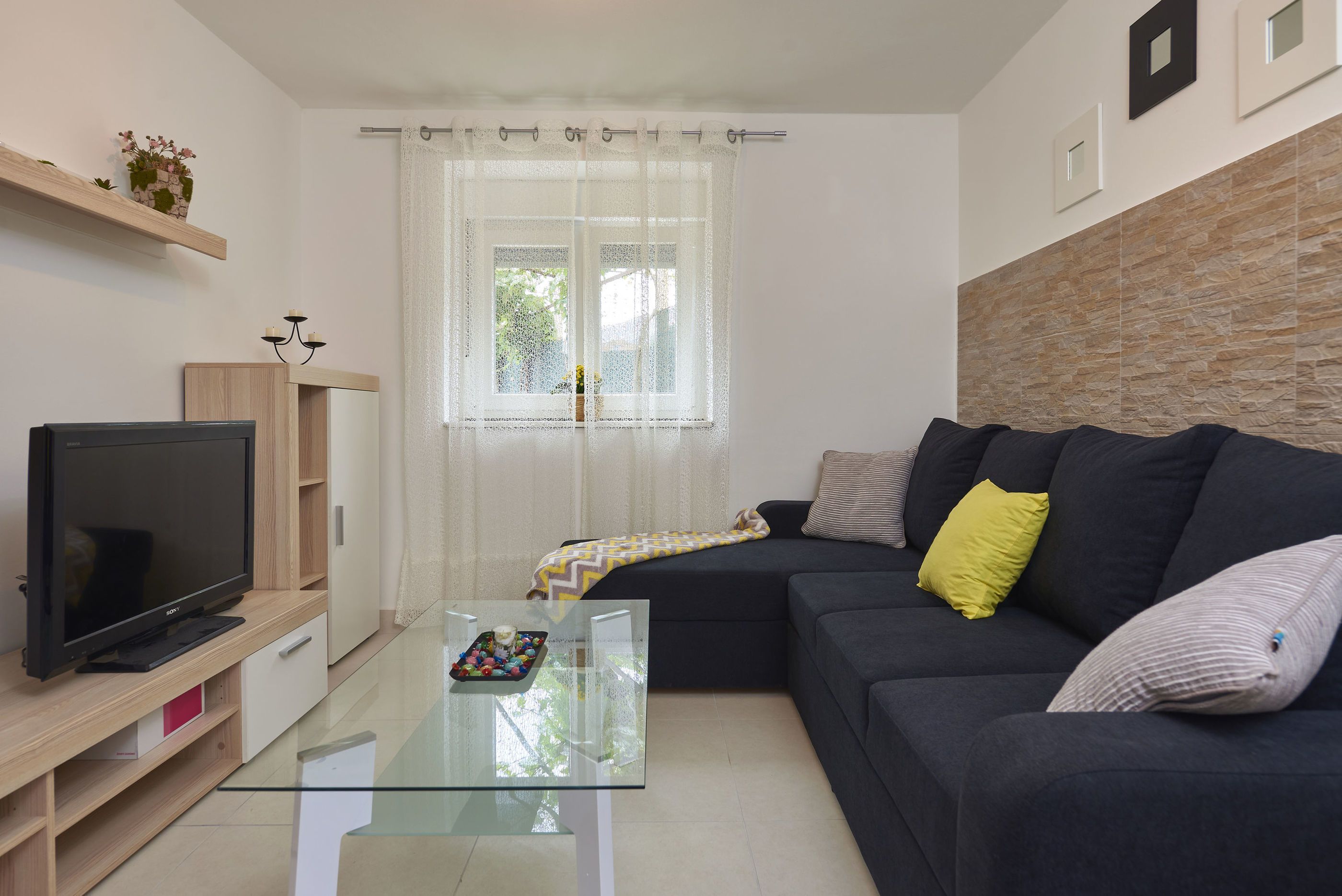 Una sala de estar moderna con un televisor de pantalla plana, una mesa de centro de vidrio, un sofá oscuro y una ventana con cortinas ligeras.