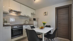 Une cuisine moderne avec des armoires blanches, un four à micro-ondes, un four traditionnel et une table à manger avec des chaises noires. Il y a une horloge au mur et une porte avec un panneau en bois.