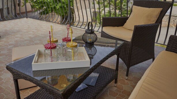 Une table à plateau en verre, sur laquelle sont disposés un plateau de bocaux et un chandelier, accompagnée de chaises en osier et d’un canapé rembourré, le tout sur un balcon doté d’une rambarde en métal.