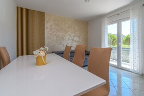 A dining area with a white table, six brown chairs, a vase with flowers, a sofa, and a large window with curtains. - Apartments Joka (Lopar)