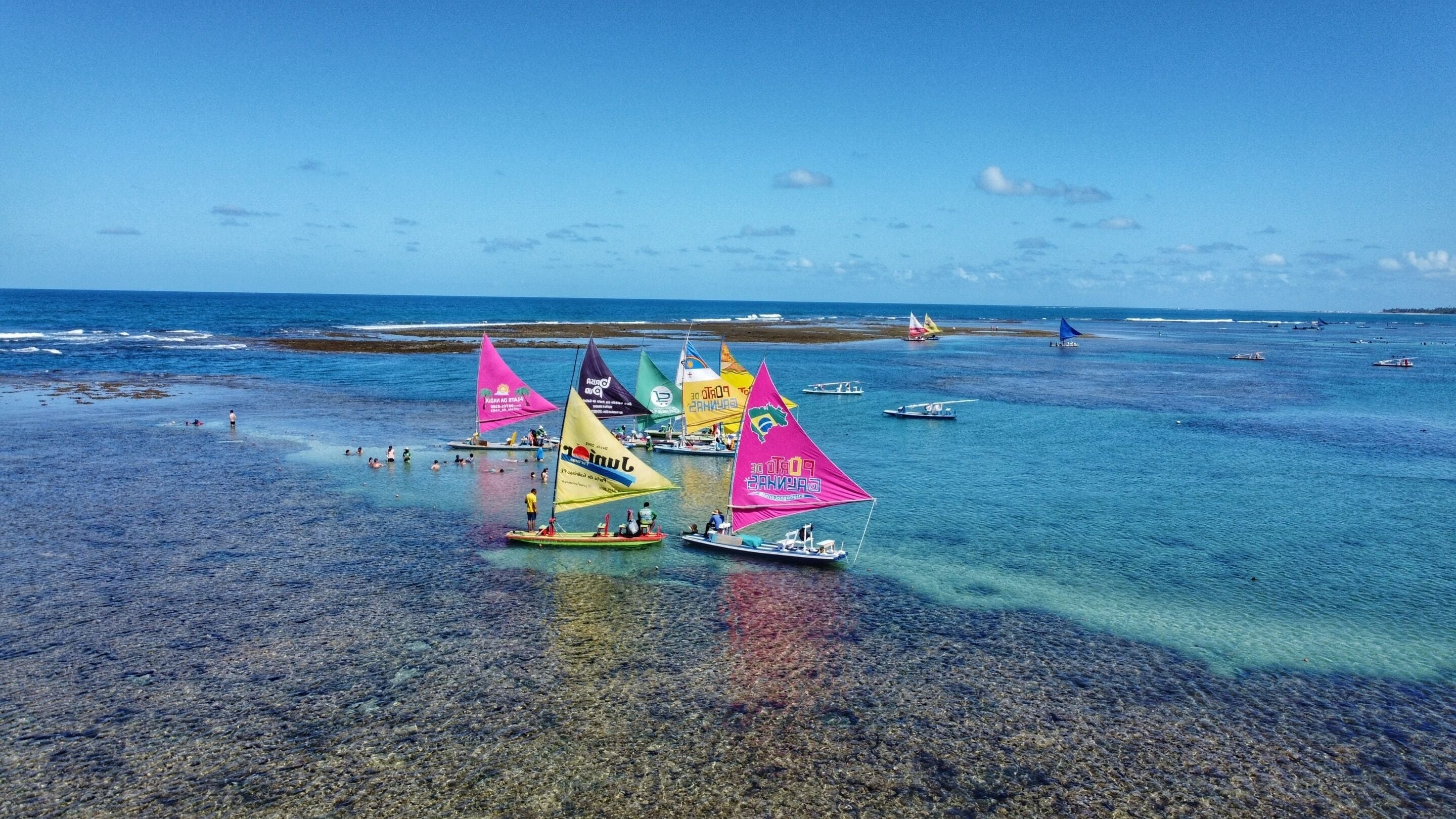 Una playa con aguas transparentes y poco profundas, barcos y una costa distante.
