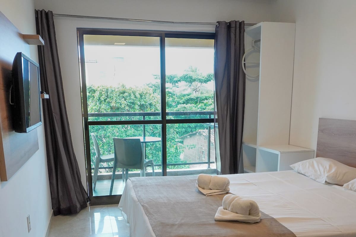 A hotel room with a bed, a TV, a balcony with a chair, and a view of greenery.