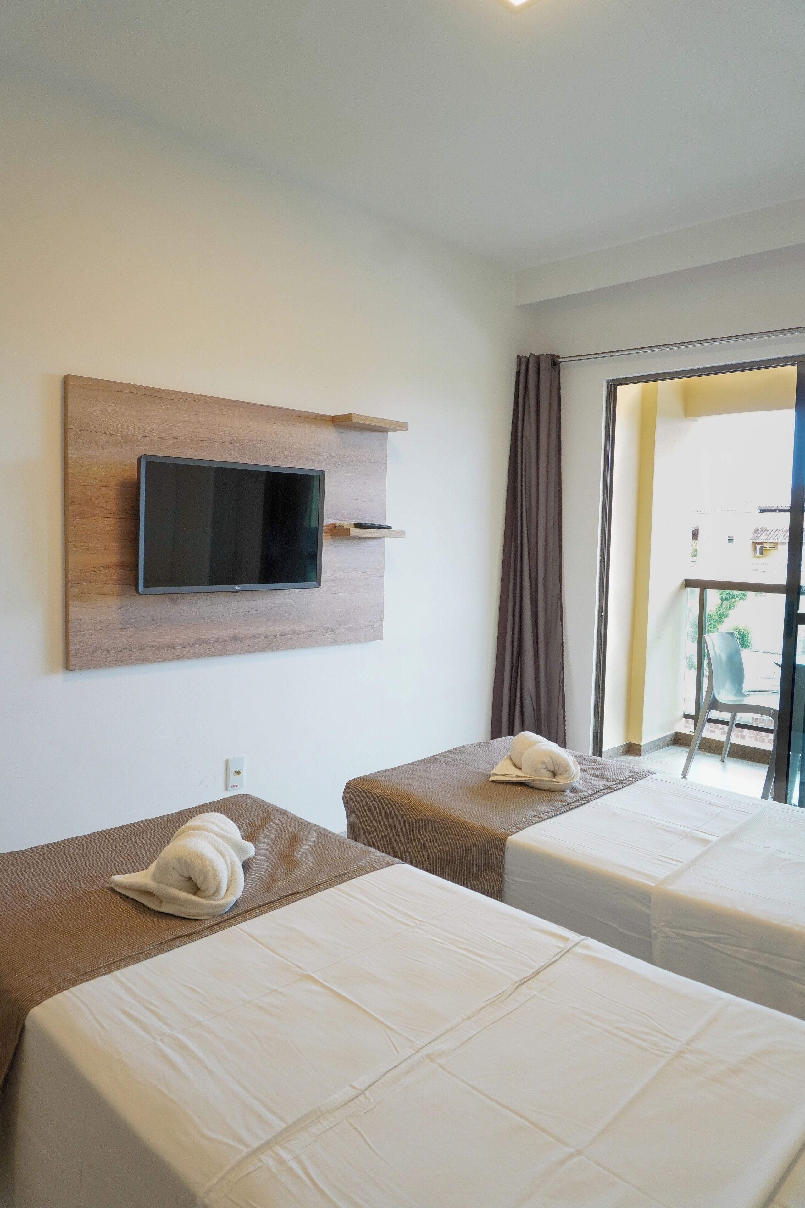 A hotel room with two beds, a mounted TV, a balcony with a chair, and a window with curtains.