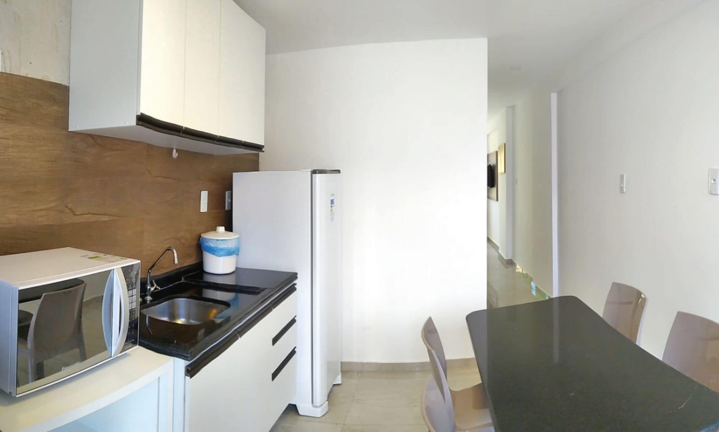 Une cuisine compacte avec des meubles blancs, un plan de travail noir, un micro-ondes encastré, un réfrigérateur blanc et une table à manger avec des chaises.