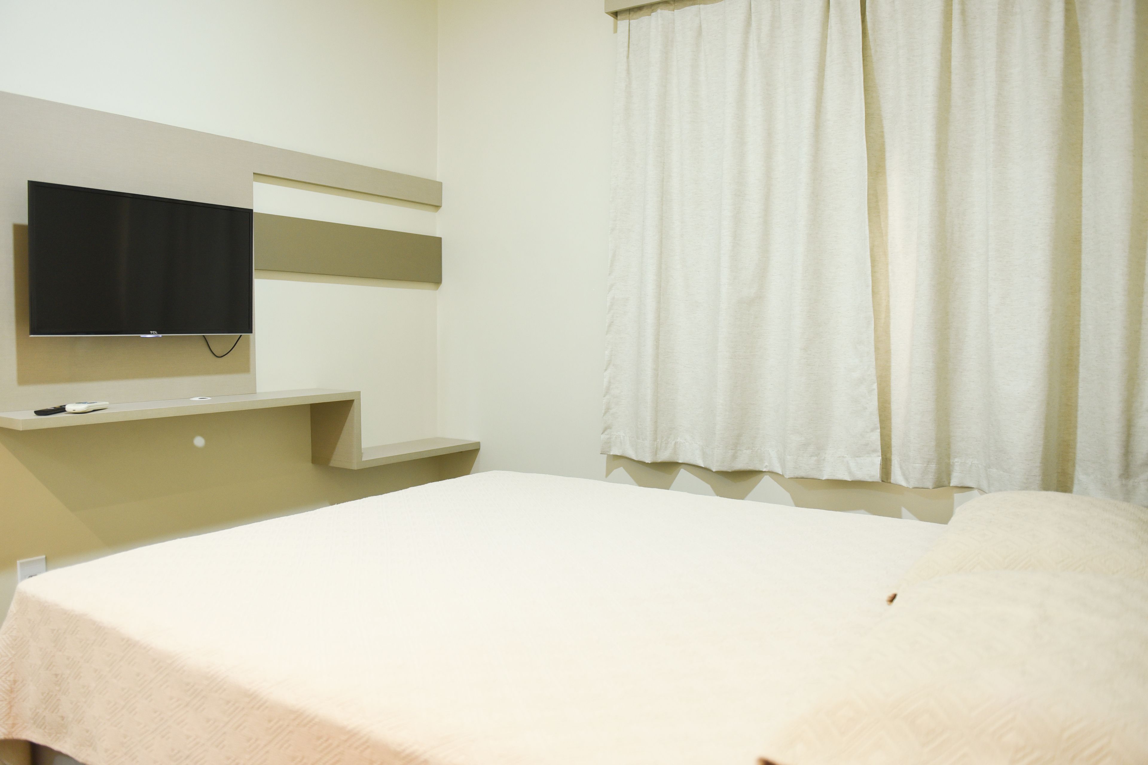 A hotel room with a bed, a flat-screen TV mounted on the wall, and a window with curtains.