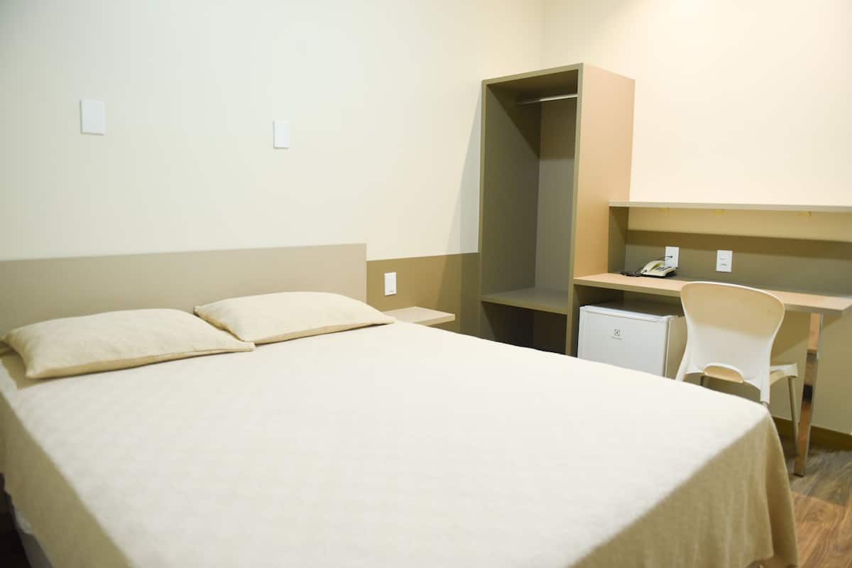 A hotel room with a bed, a desk, a chair, and a small refrigerator.