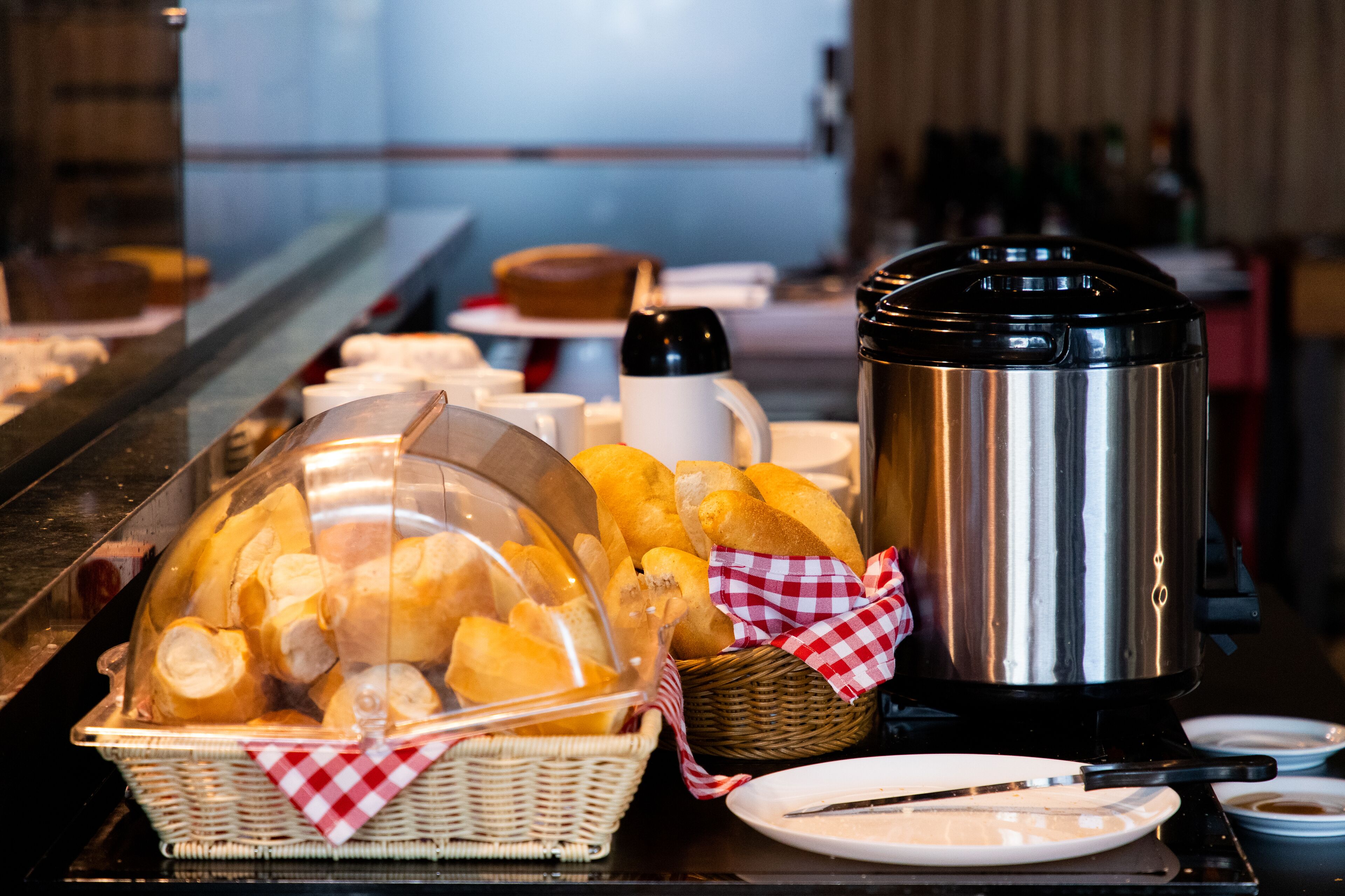 A buffet setup with a basket of bread, a coffee pot, and plates with utensils.