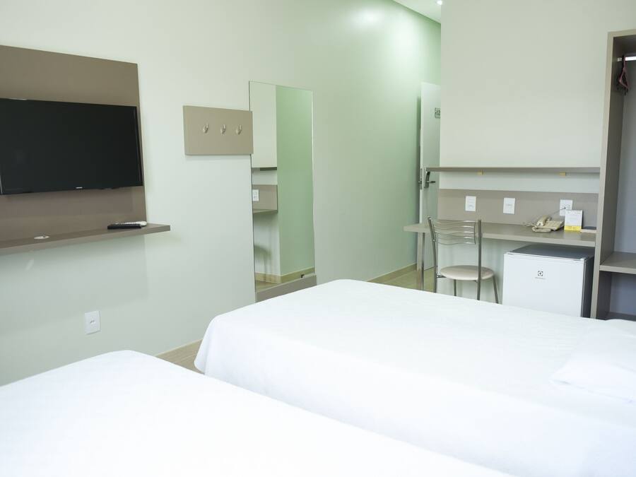 Habitación de hotel con dos camas, televisión, escritorio con silla, espejo y refrigerador pequeño.