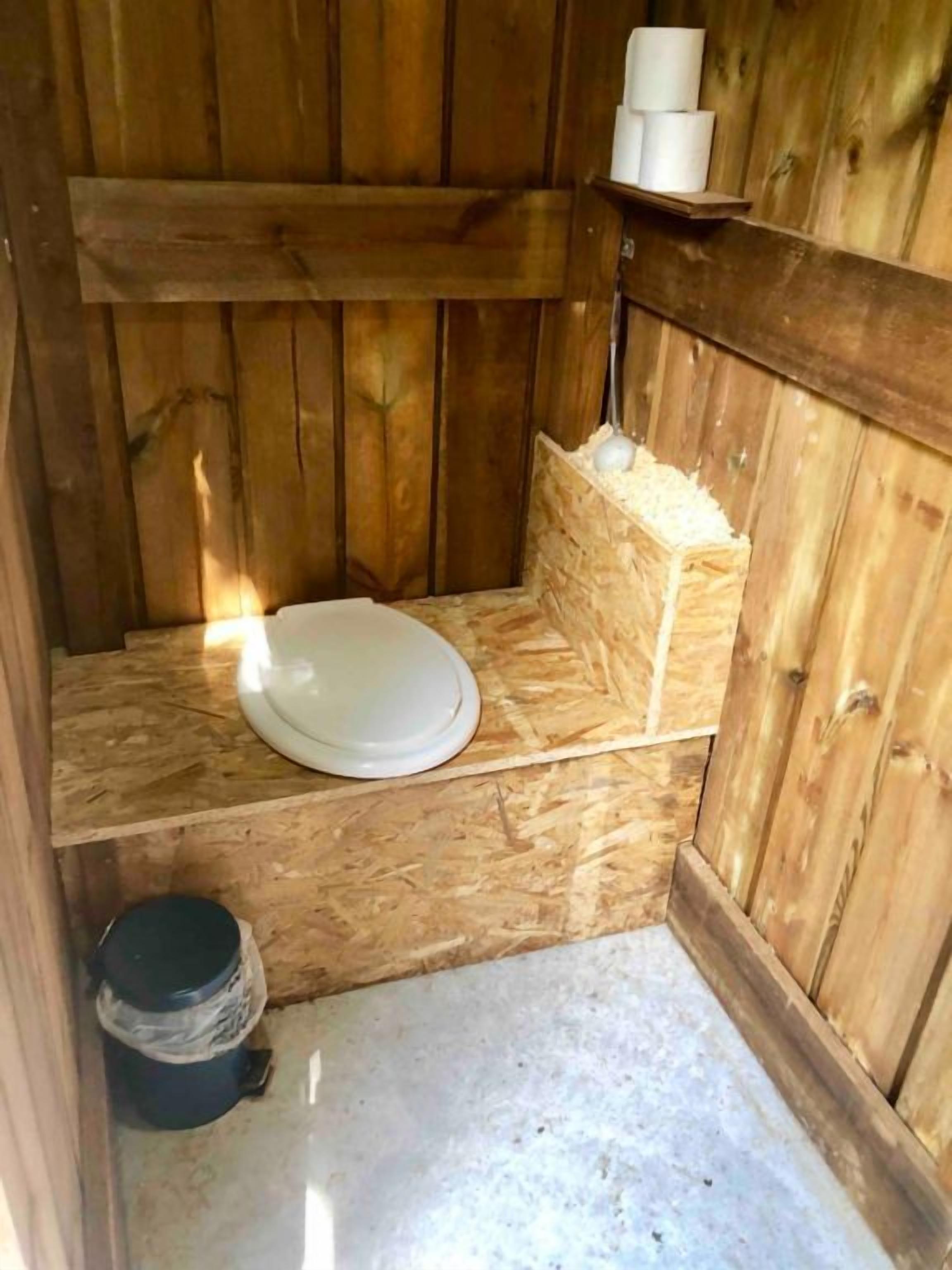 A small wooden cabin with a single toilet, a shelf with toilet paper, and a trash bin.