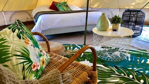 Een gezellig ingerichte tent met een bed, rieten meubels en een klein rond tafeltje met een plant.