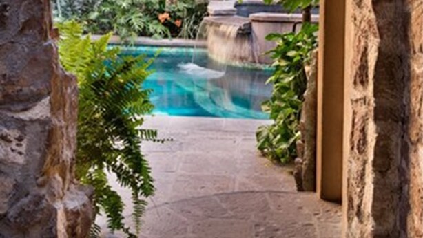 An archway leading to a pool area with a waterfall, surrounded by lush greenery and stone walls.