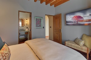 A bedroom with a bed, a bench with cushions, a painting, a mirror, and a door leading to a bathroom.