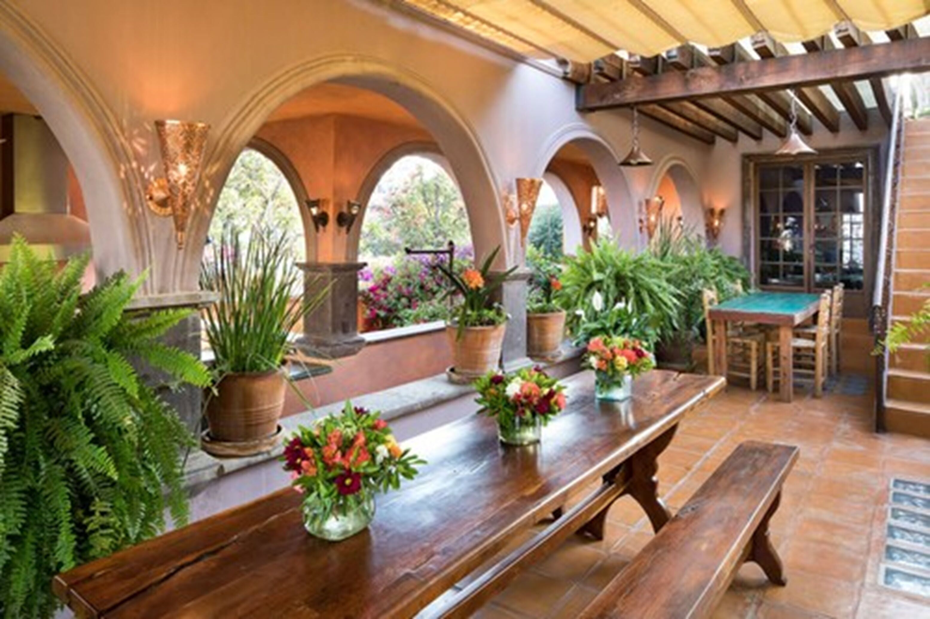 A covered patio with a wooden table, benches, and potted plants.