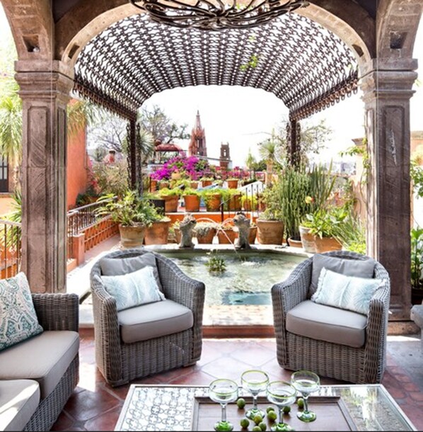 A patio area with wicker furniture and a glass table, overlooking a garden with a fountain and potted plants.
