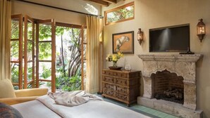 A bedroom with a bed, wooden ceiling beams, a fireplace, a television, and a window with curtains.