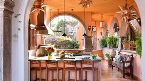 An interior with a stone archway leading to a kitchen area featuring a central island with stools, overhead lighting fixtures, and potted plants.