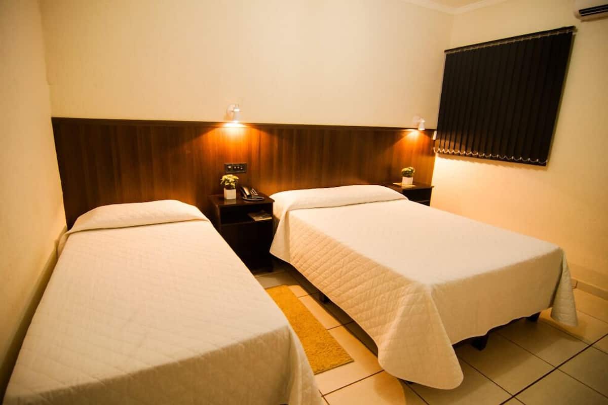 Une chambre d’hôtel avec deux lits simples, une tête de lit en bois, des tables de chevet avec des lampes et une fenêtre avec des stores.