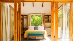A bedroom with a bed, wooden beams, a window with curtains, and a view of greenery outside.