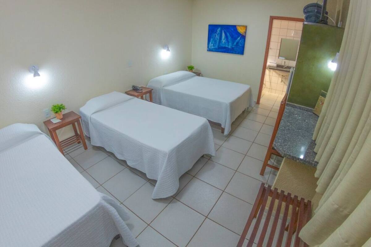 A room with three beds, a small table with a plant, a painting on the wall, and a bathroom visible through an open door.