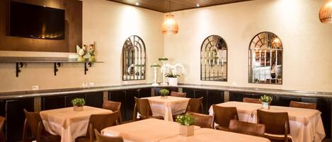 A restaurant with multiple tables and chairs, a television mounted on the wall, and decorative plants.