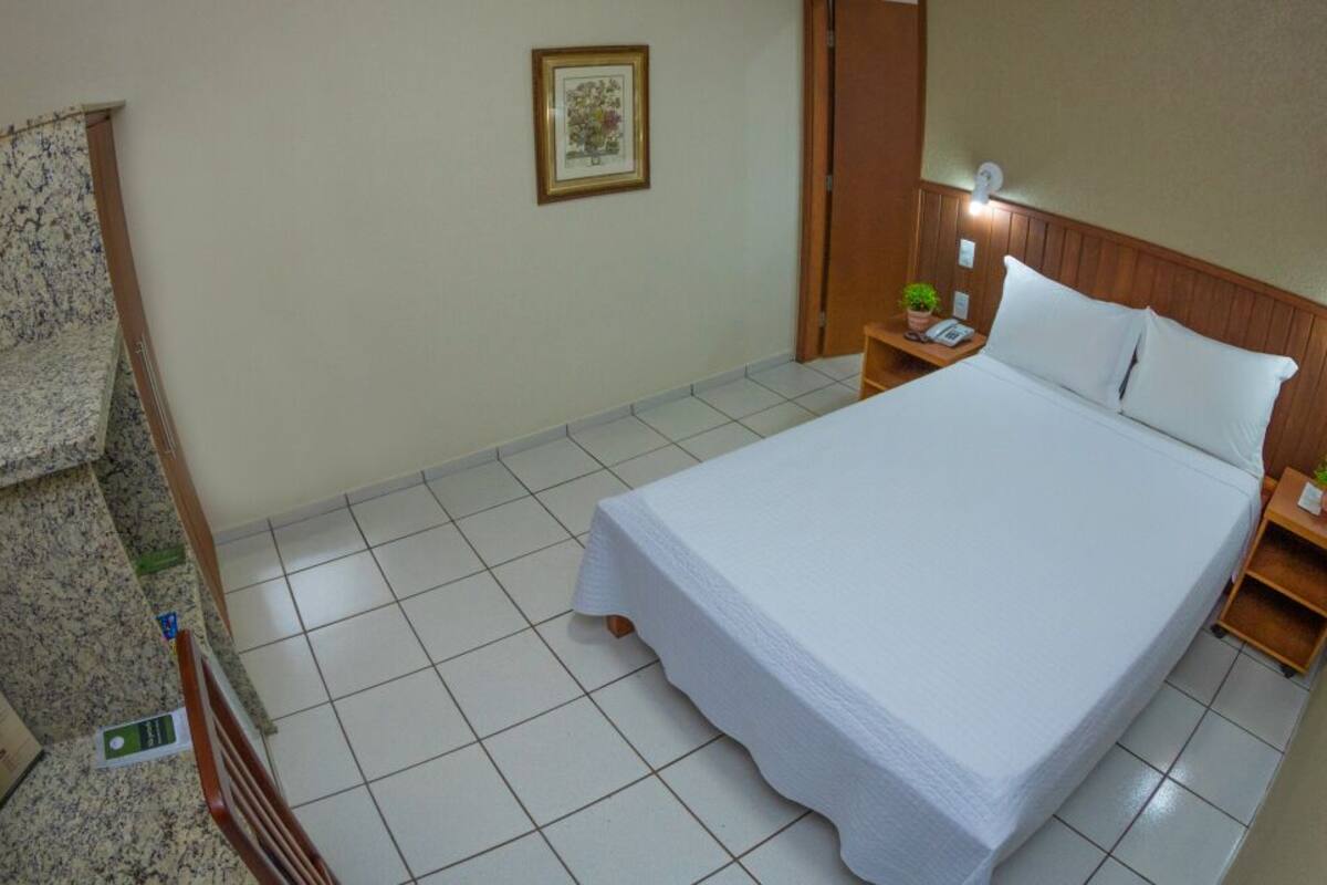 A hotel room with a large bed, a wooden headboard, a nightstand with a lamp, a small table with a plant, and a framed picture on the wall.