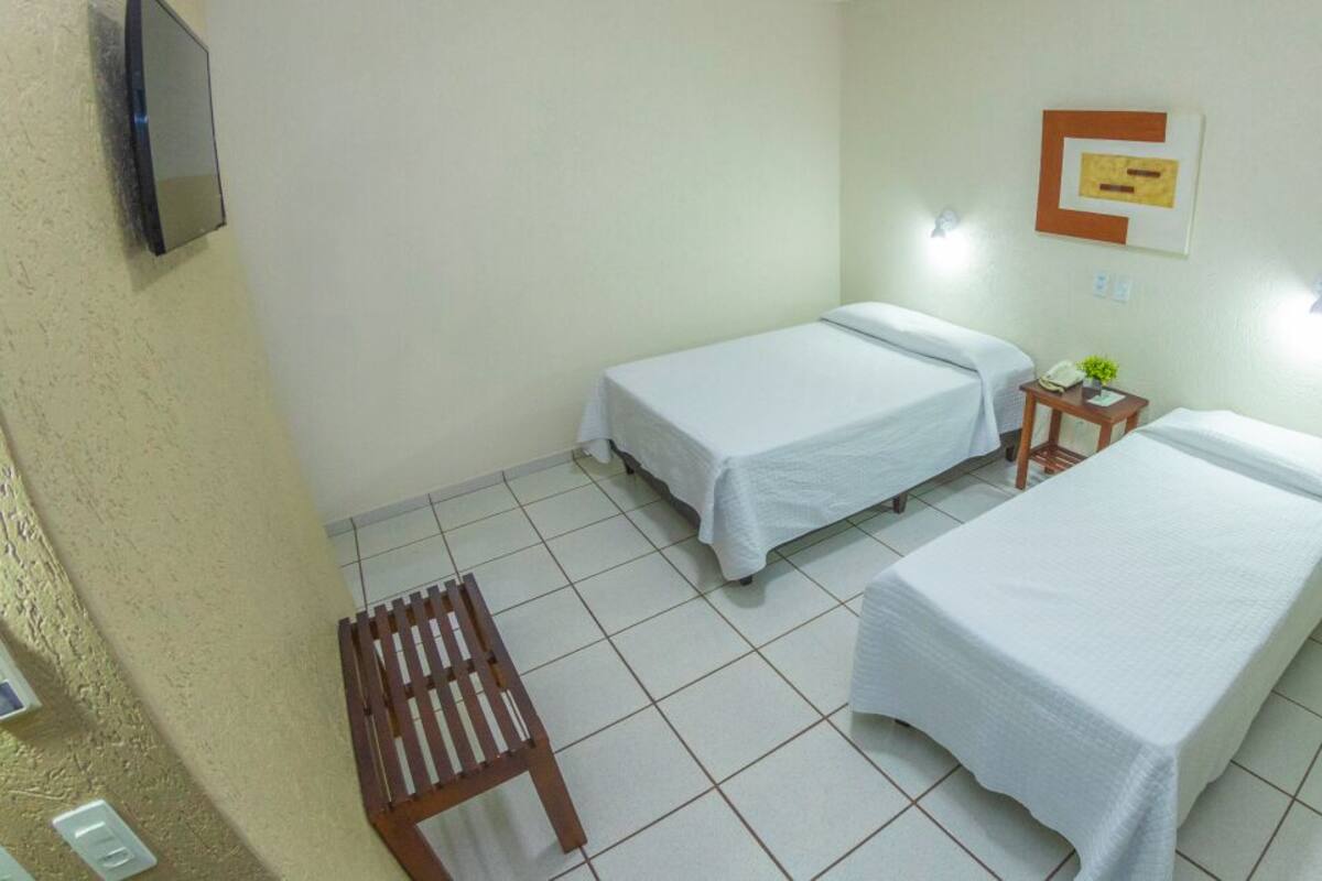 A room with two single beds, a small table with a lamp, and a wall-mounted TV.