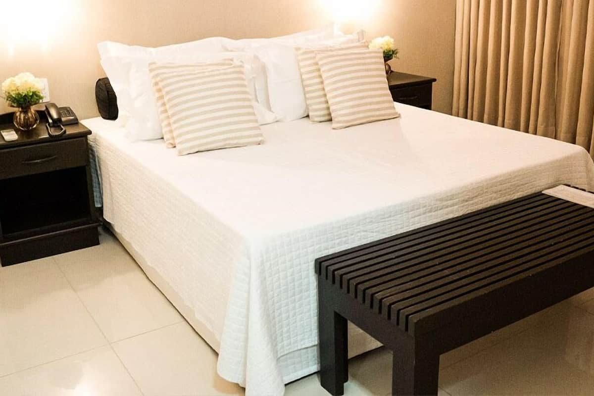 A neatly made bed with white bedding and striped pillows, a bedside table with a phone and a vase of flowers, and a wooden bench in front of the bed.