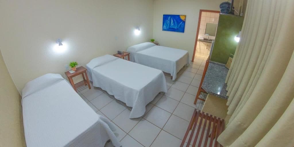 Quarto de hotel com três camas de solteiro, uma mesinha com uma planta, um quadro na parede e um banheiro visível através de uma porta aberta.