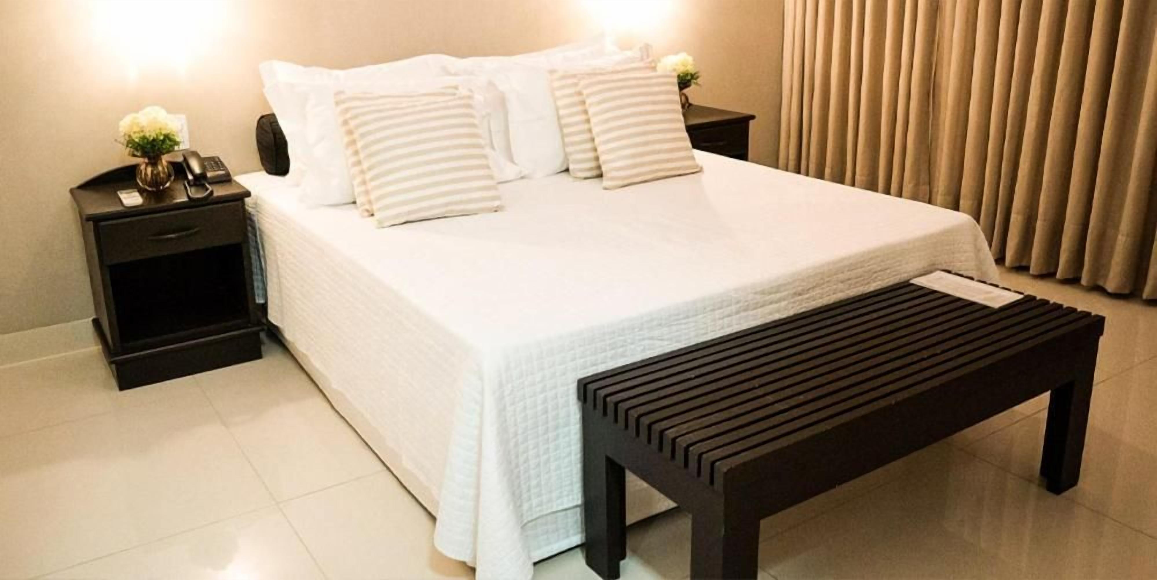 Una cama bien hecha con ropa de cama blanca y almohadas a rayas, una mesita de noche con una lámpara y un teléfono, y un banco de madera frente a la cama.