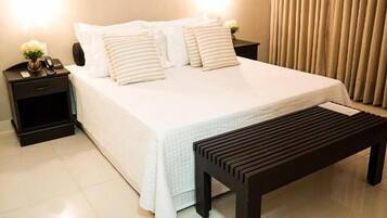 Una cama bien hecha con ropa de cama blanca y almohadas a rayas, una mesita de noche con una lámpara y un teléfono, y un banco de madera frente a la cama.