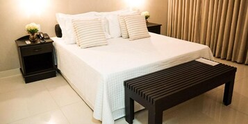 Una cama bien hecha con ropa de cama blanca y almohadas a rayas, una mesita de noche con una lámpara y un teléfono, y un banco de madera frente a la cama.
