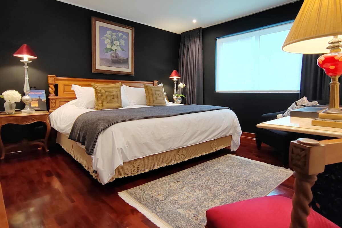 A hotel room with a large bed, a wooden headboard, a nightstand with a lamp, a desk with a lamp, a window with curtains, and a framed picture on the wall.