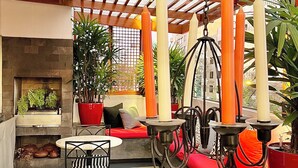 Un área de patio al aire libre con una pérgola de madera, un espacio de descanso con cojines rojos y una mesa con sillas. Hay plantas en macetas y una lámpara colgante.