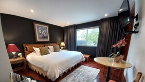 Una habitación de hotel con una cama grande, mesitas de noche, una mesa redonda, un sofá y un televisor montado en la pared.