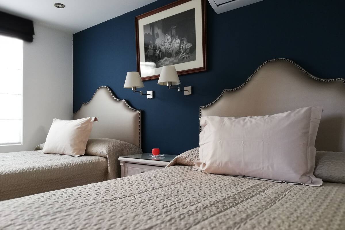 A hotel room with a large bed, two smaller beds, a nightstand with a lamp, a framed picture on the wall, and a window with curtains.