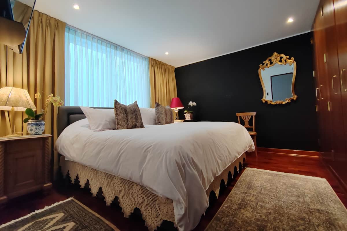 A bedroom with a large bed, a nightstand with a lamp, a potted plant, a chair, and a mirror on the wall.