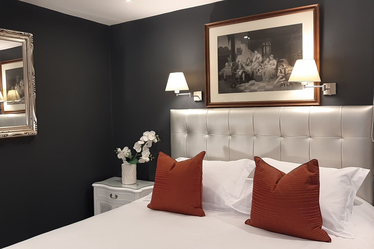 A bedroom with a large bed, a nightstand with a vase of flowers, a framed picture above the bed, and a mirror on the wall.