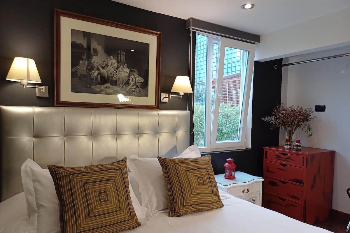 A hotel room with a tufted headboard, a large framed black and white photograph, two bedside lamps, a window with a view of buildings, a red dresser, and a vase with flowers.