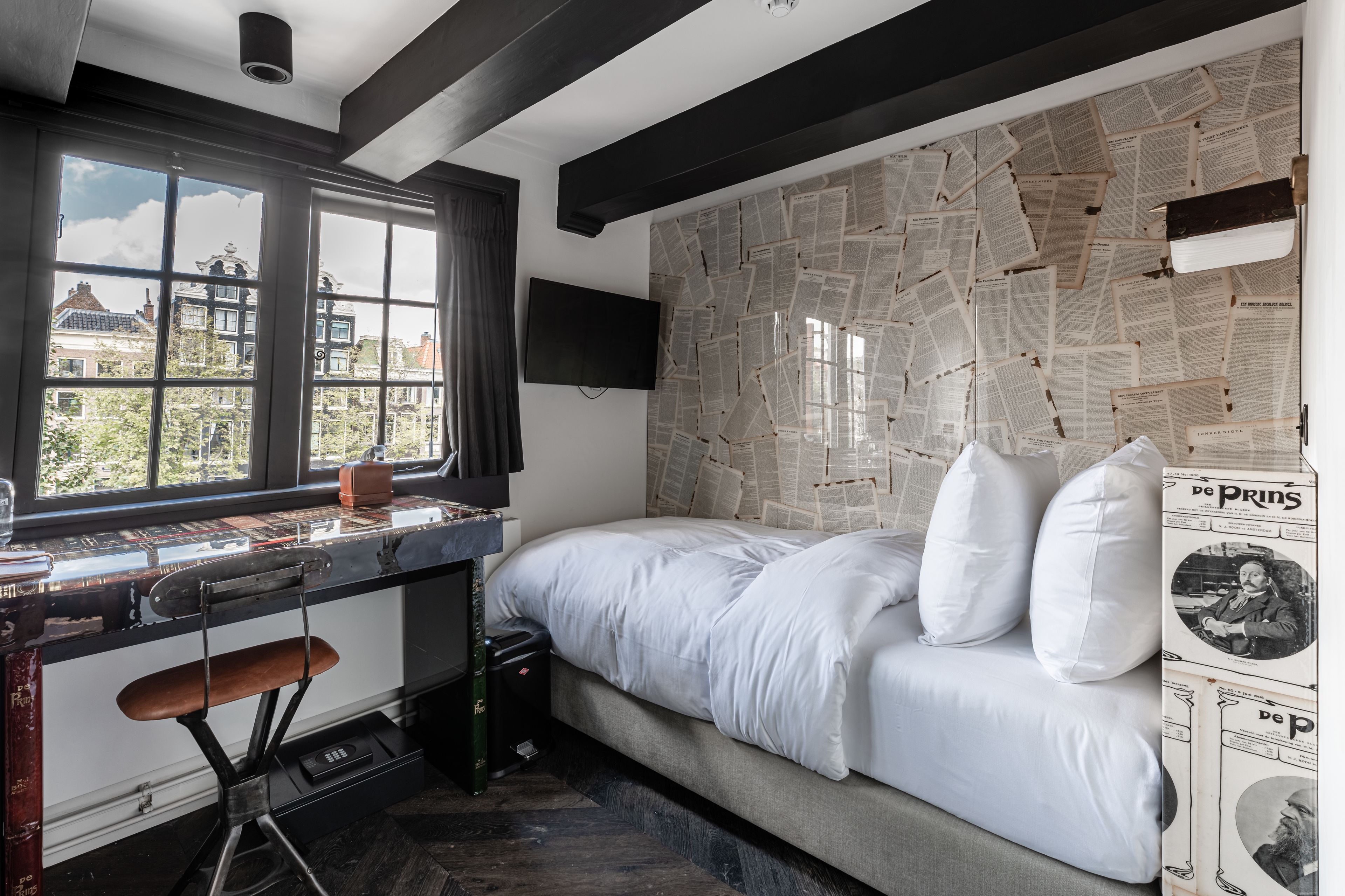 A bedroom with a bed, a desk, a chair, and a window with a view of buildings.