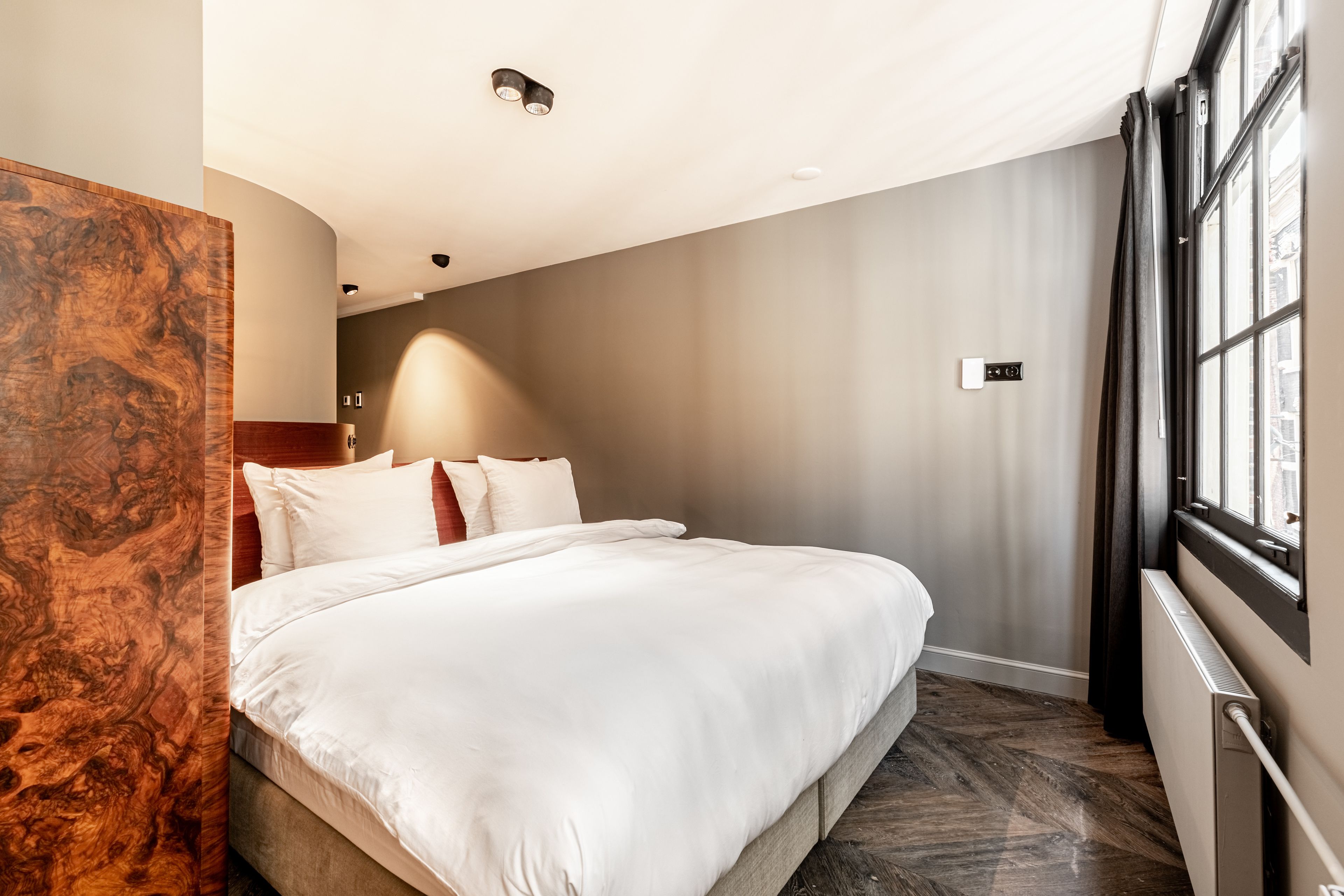 A modern hotel room with a large bed, a wooden accent wall, a built-in heater, and a window with curtains.